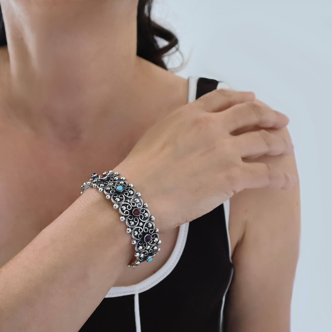 A close-up of a woman wearing a wide Mexican 925 sterling silver filigree bracelet with small turquoise blue stones, amethyst and dark coral red accents on her wrist.