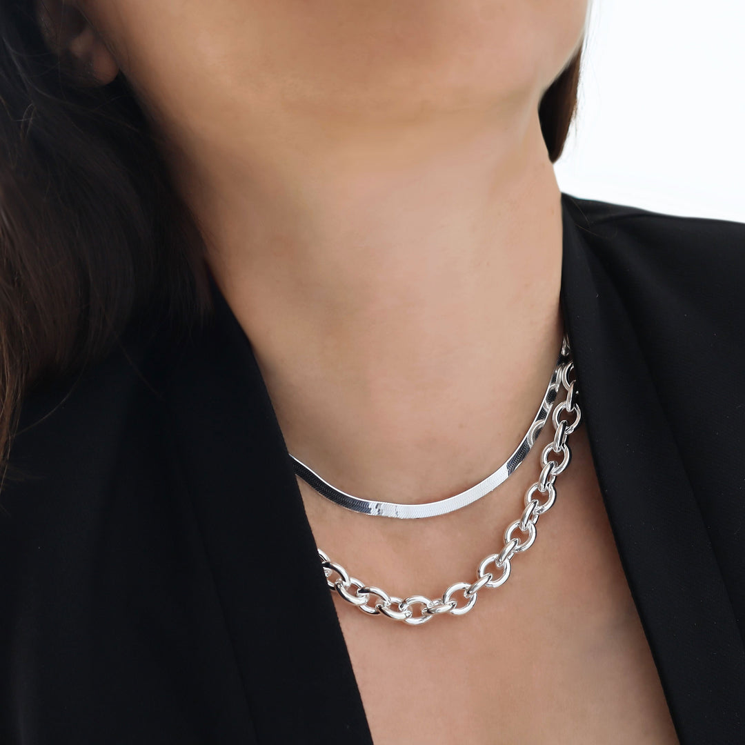 Silver chunky chain necklace and flat herringbone chain necklace stack worn by a woman with a black jacket. 