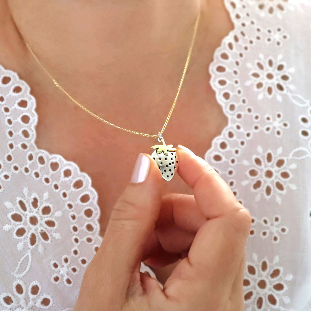 Necklace with a strawberry pendant and gold chain held by a hand against a white lace background