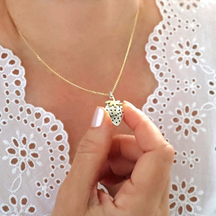 Necklace with a strawberry pendant and gold chain held by a hand against a white lace background