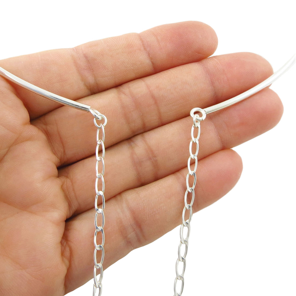 A close-up of a hand holding a thin silver choker torque necklace with two dangling chain extensions against a plain white background.