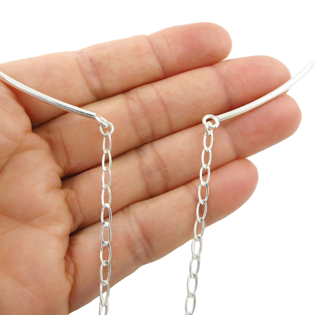 A close-up of a hand holding a thin silver choker torque necklace with two dangling chain extensions against a plain white background.
