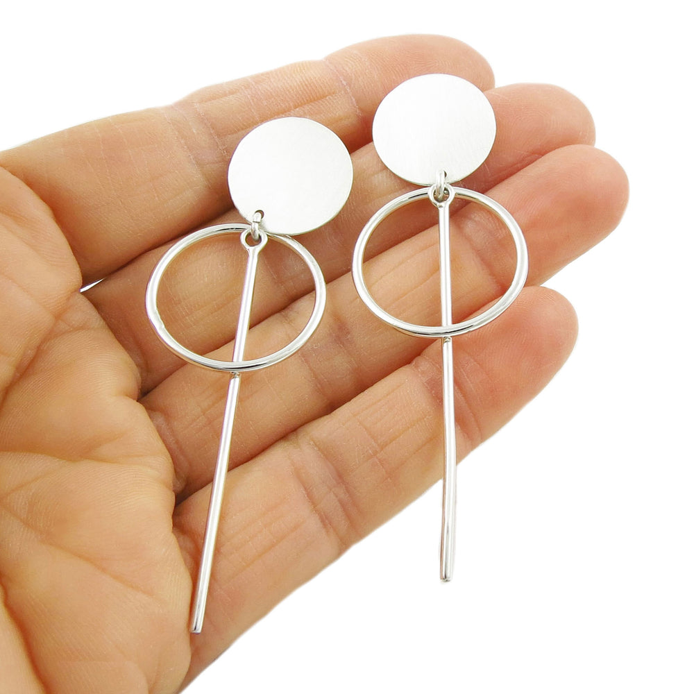 A pair of dangling sterling silver earrings with a satin finish held in a hand, displaying the elegant drop design with long slender sticks and circular discs.