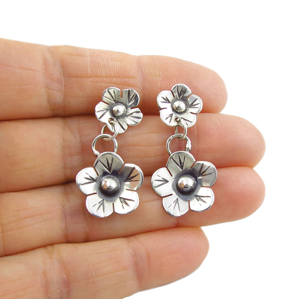 A pair of silver flower drop earrings held between fingers, displaying their dangle and detailed flower design.