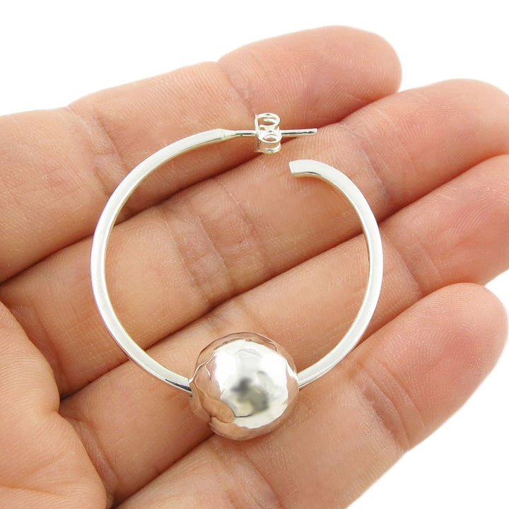 A single 925 silver hoop earring with a spherical hammered ball bead charm held in a hand on a white background