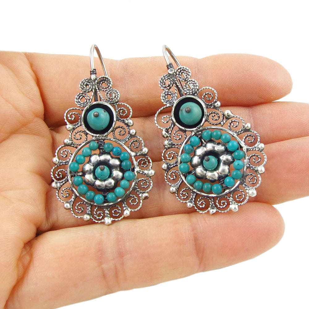 Ornate silver and turquoise earrings held in a hand against a white background