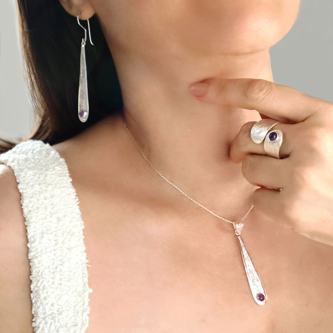 Close-up of a woman wearing a silver necklace with a pendant, ring, and matching earrings with a crumpled reticulated finish and amethyst gemstones. 