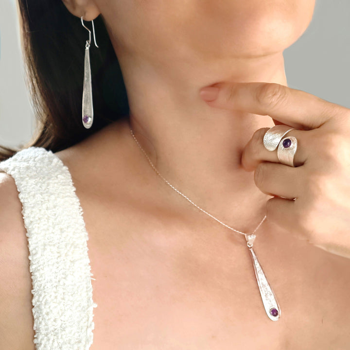 Close-up of a woman wearing a silver necklace with a pendant, ring, and matching earrings with a crumpled reticulated finish and amethyst gemstones. 