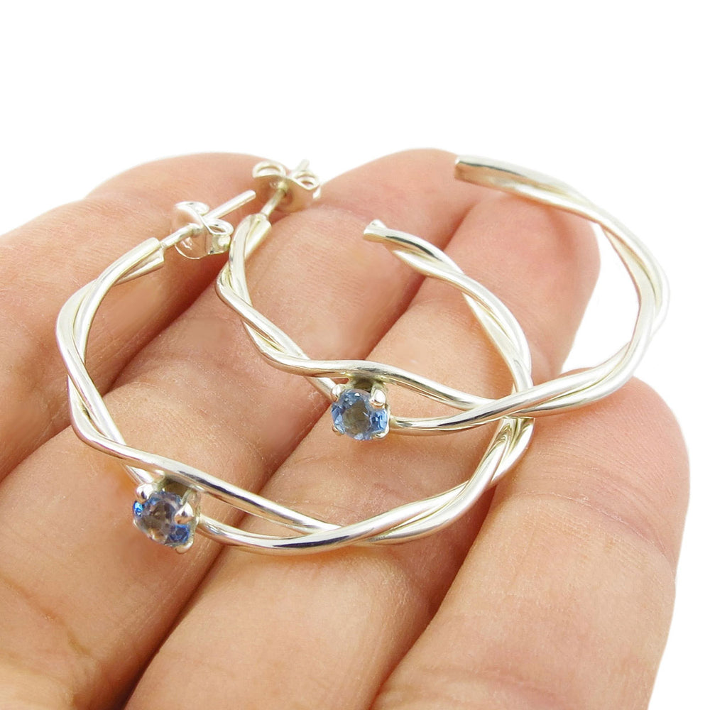 Silver hoop earrings with blue topaz  gemstones held in a hand on a white background