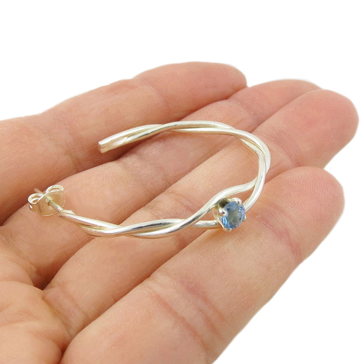A single silver hoop earring with a blue topaz gemstone on a hand against a white background
