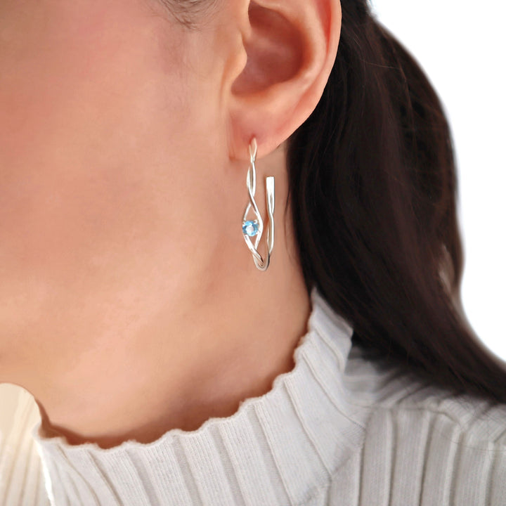 Silver earrings with a faceted blue topaz gemstone worn by a person against a neutral background