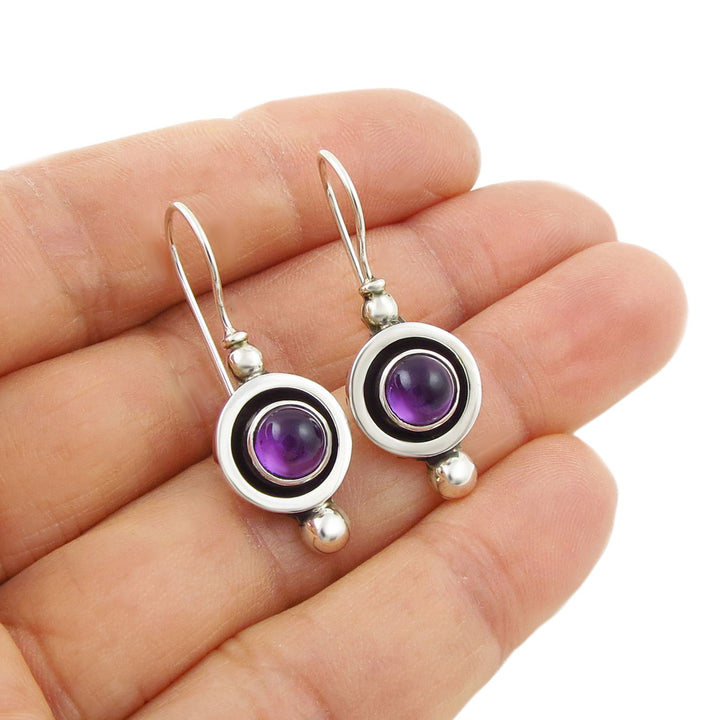 Silver earrings with purple amethyst gemstones held in a hand on a white background