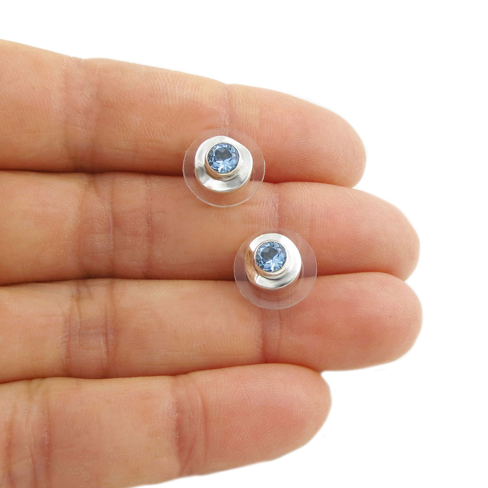 Two small silver earrings with blue topaz gemstones held between fingers on a white background