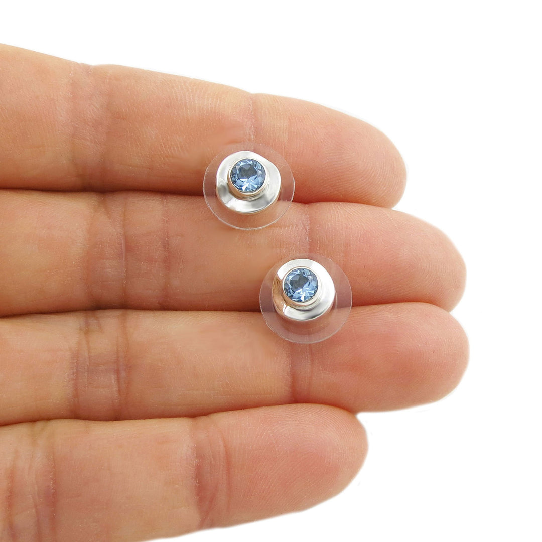 Two small silver earrings with blue topaz gemstones held between fingers on a white background