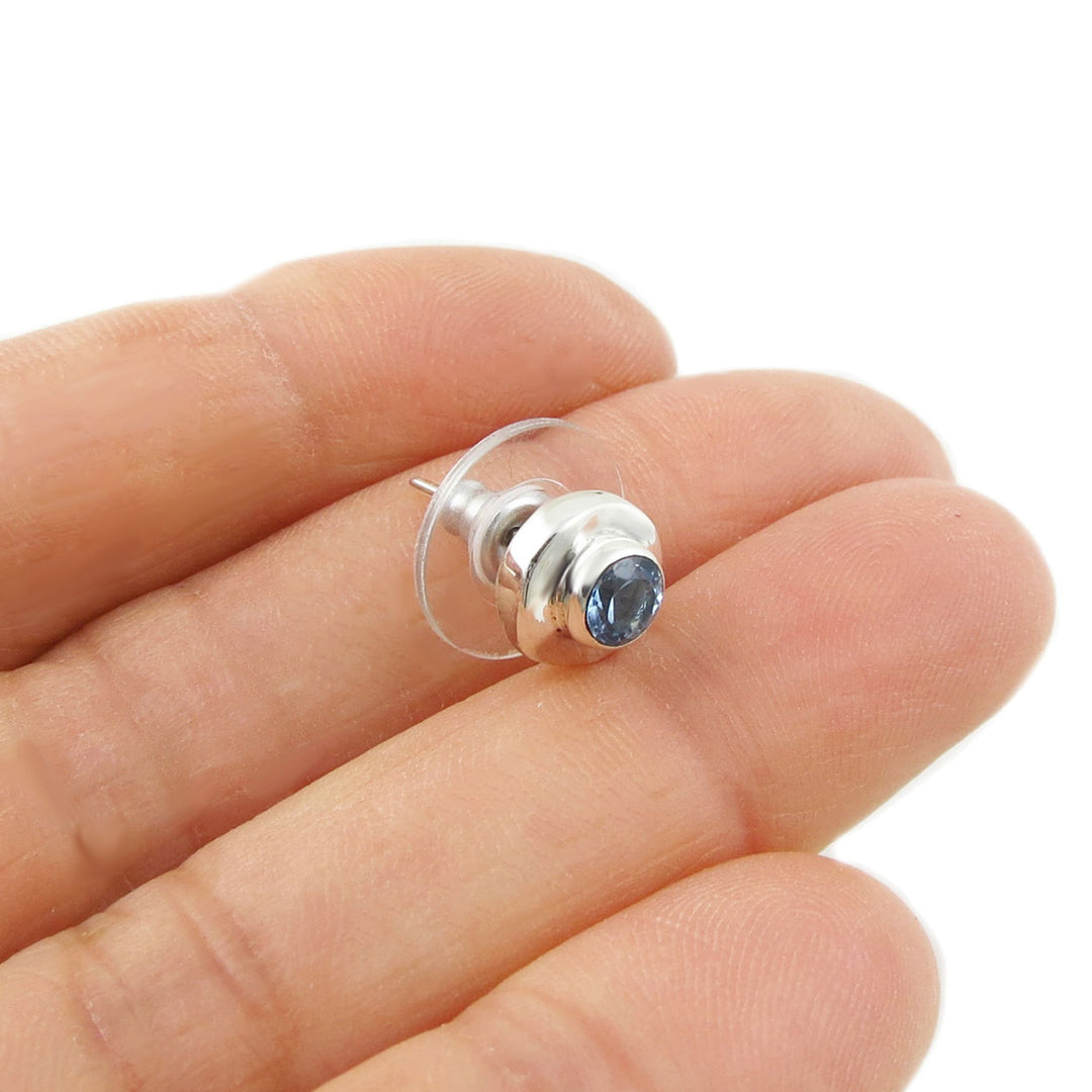 A single silver earring with a blue topaz gemstone on a hand against a white background