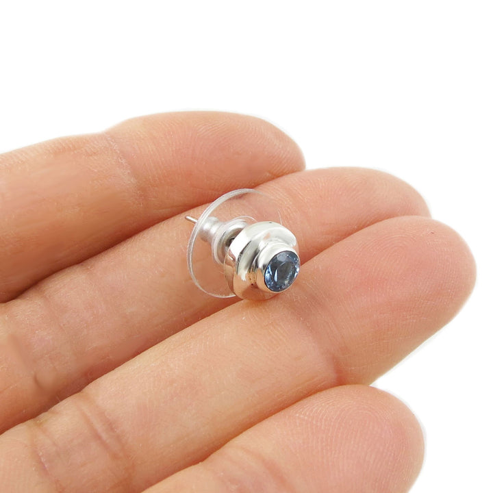 A single silver earring with a blue topaz gemstone on a hand against a white background