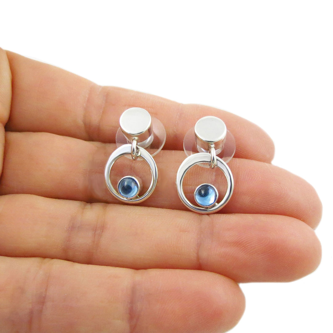 Silver earrings with blue topaz gemstones held between fingers on a white background