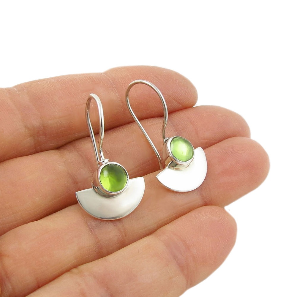 Silver earrings with green peridot gemstones held between fingers on a white background