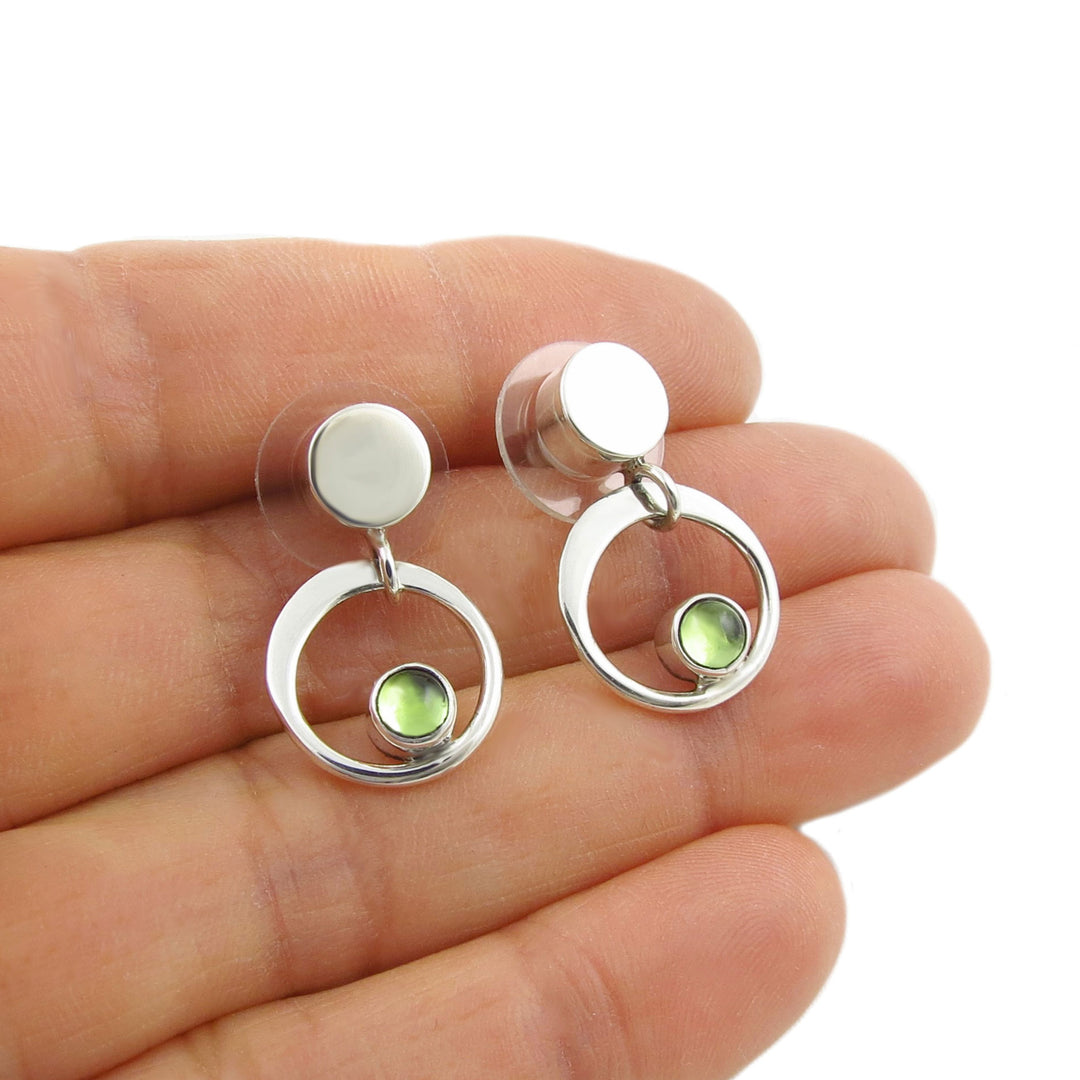 Silver earrings with green peridot gemstones held between fingers on a white background