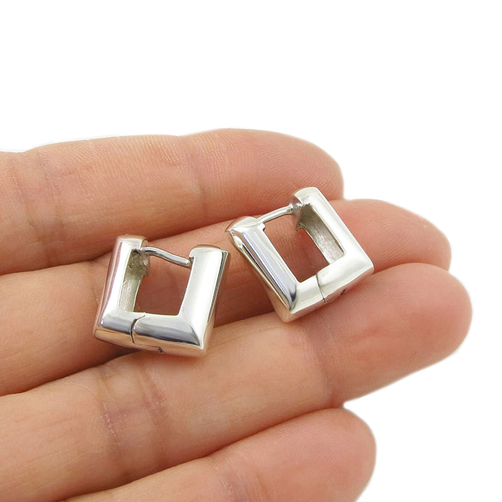 Silver square huggie hoop earrings held between fingers on a white background
