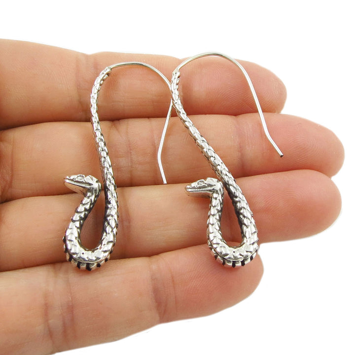 Silver snake-shaped earrings held between fingers on a white background