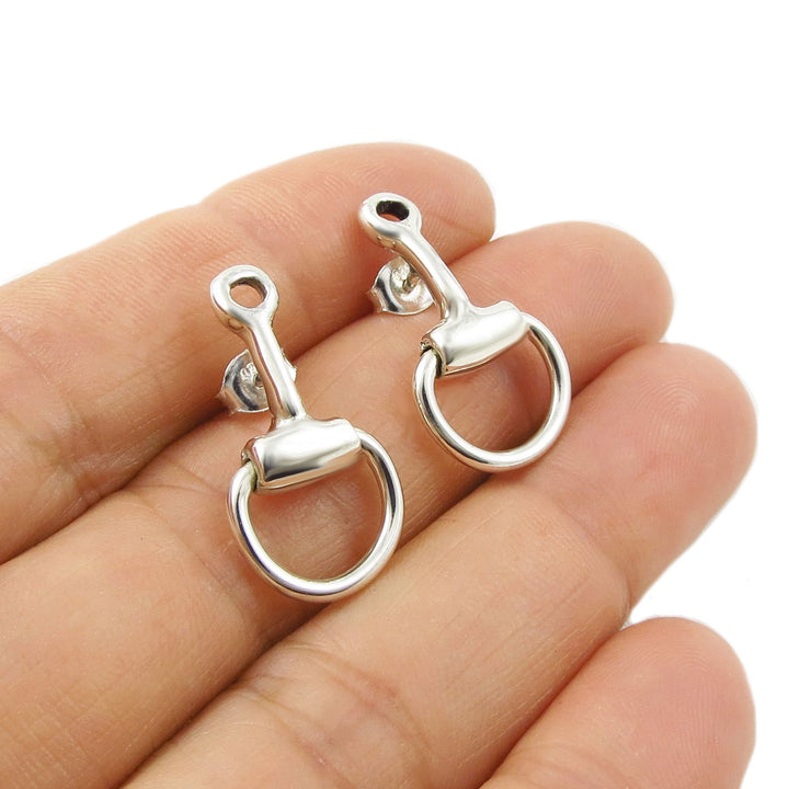 Silver equestrian-themed snaffle earrings held between fingers on a white background