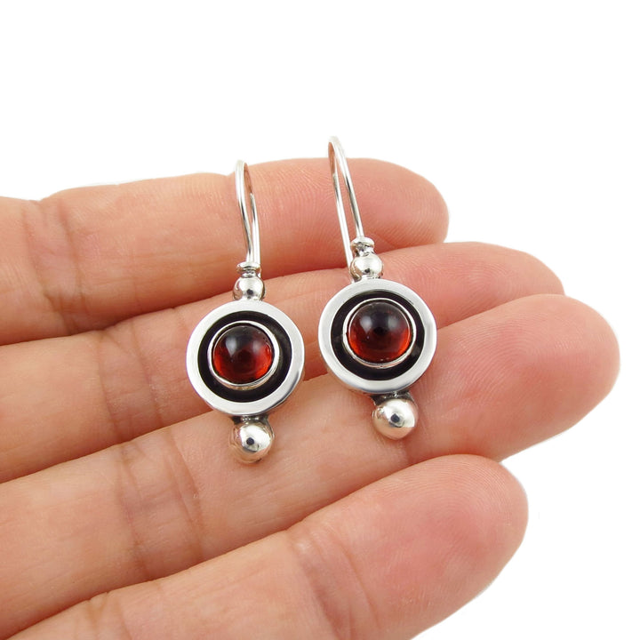 Silver earrings with red garnet gemstones held between fingers on a white background