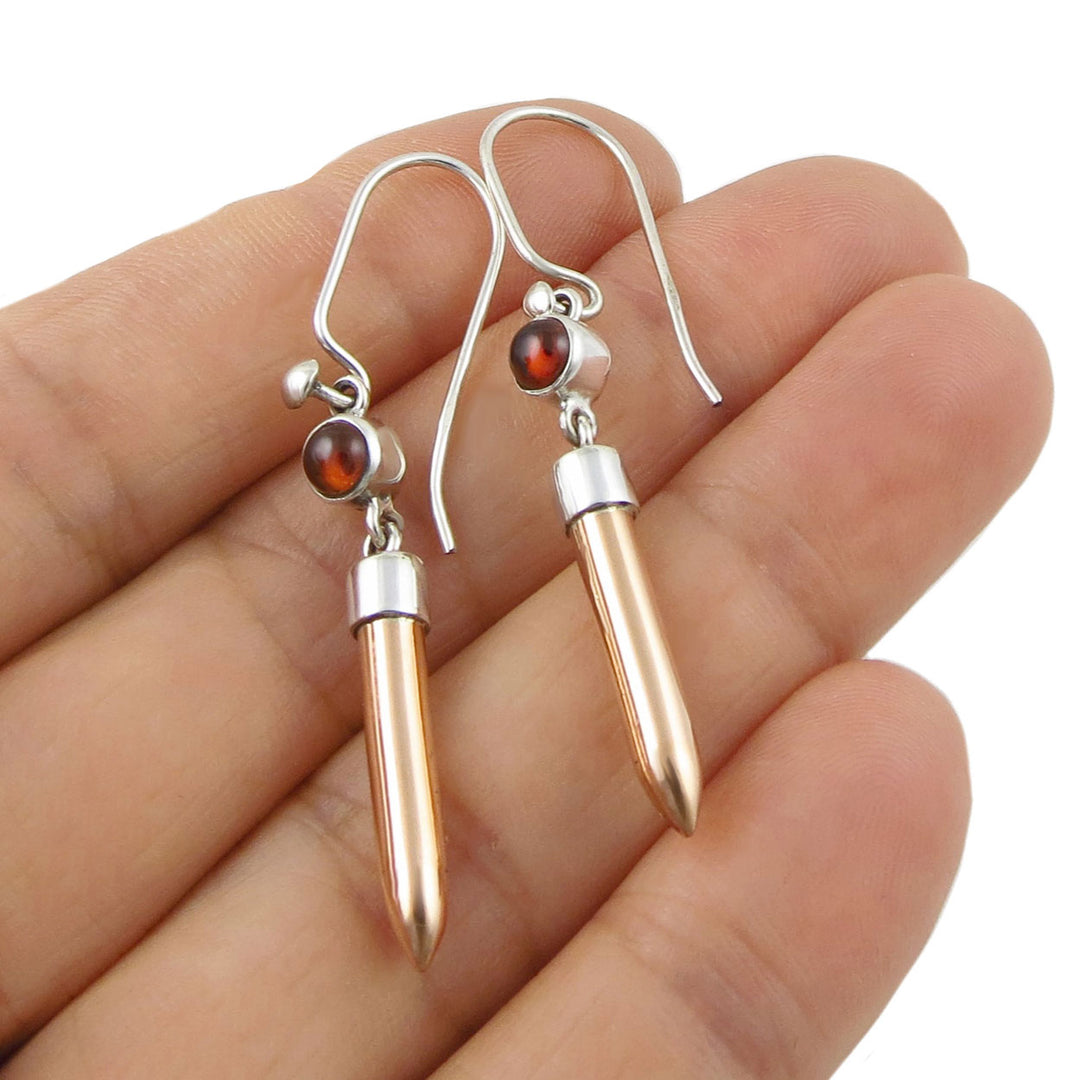 A mixed metal copper and silver drop earrings with a garnet gemstone held in a person's hand. 