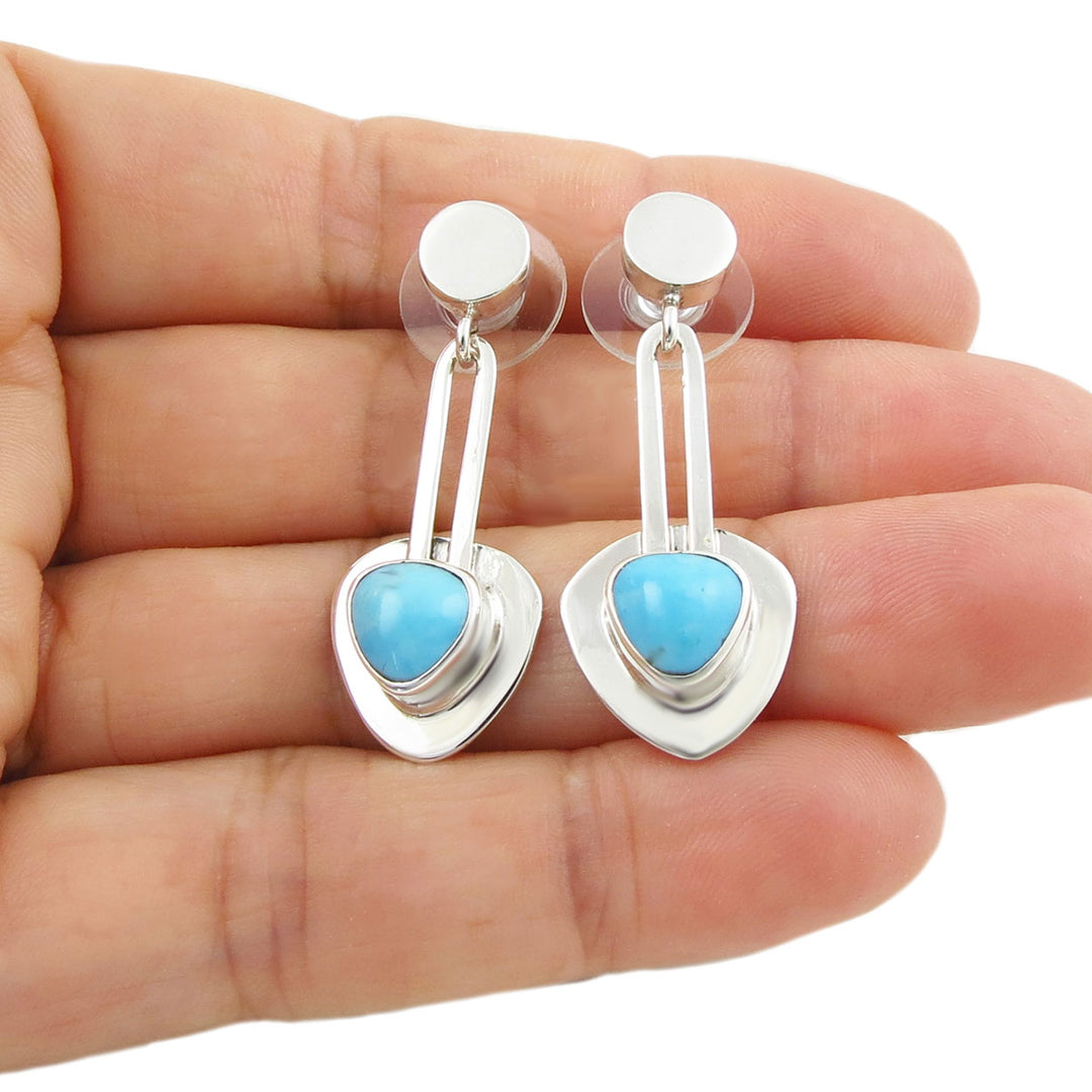 Silver earrings with blue heart-shaped turquoise gemstones held between fingers on a white background