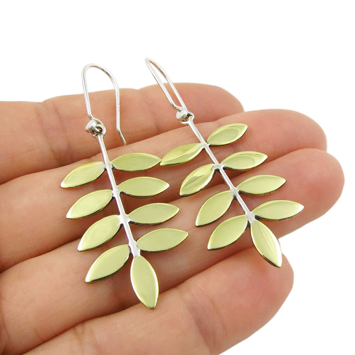 Leaf-shaped earrings in silver and gold held in a hand against a white background