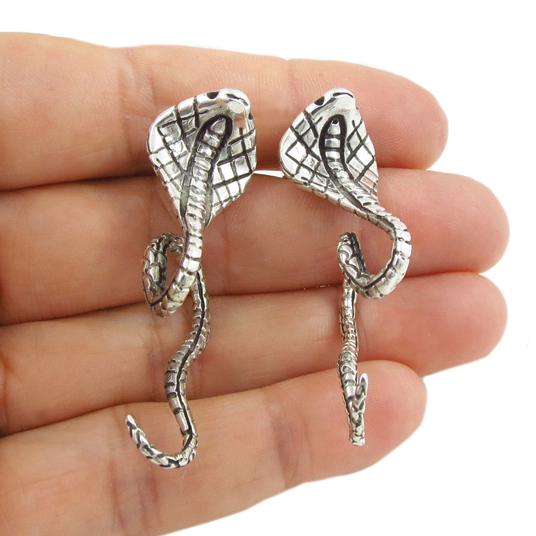 Large silver cobra snake-shaped earrings held between fingers on a white background