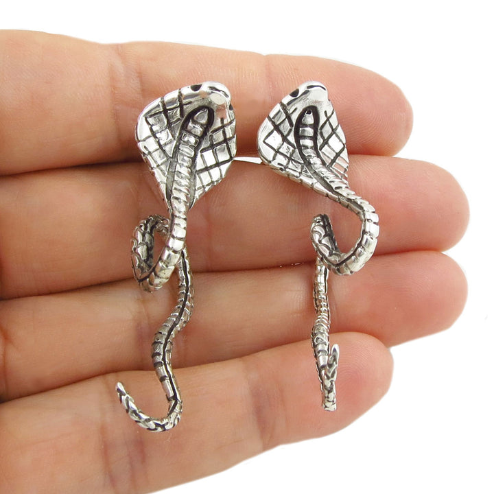 Large silver cobra snake-shaped earrings held between fingers on a white background