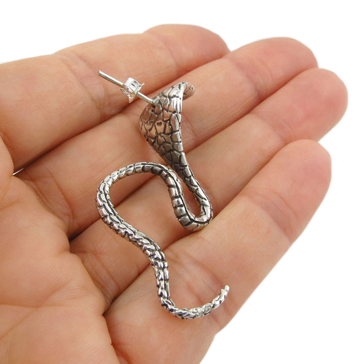 A single silver cobra snake earring held in a hand against a white background
