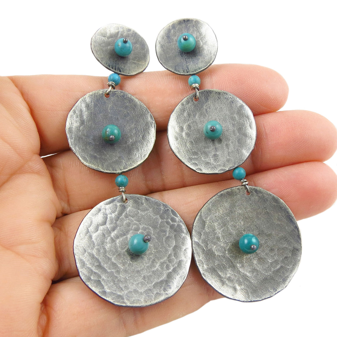 A pair of long Mexican Maria Belen Nilson chadelier earrings in a matt modern finish with turquoise accents held in a person's hand