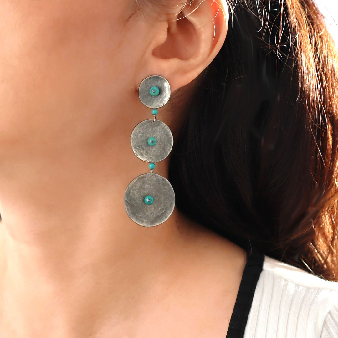 A long triple circle drop chandelier style earring in modern blackened silver with turquoise bead accents worn by  a woman with dark hair