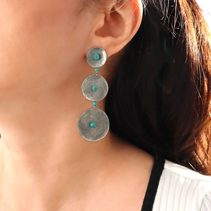A long triple circle drop chandelier style earring in modern blackened silver with turquoise bead accents worn by  a woman with dark hair