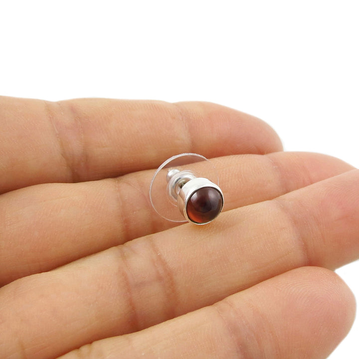 A single small round 925 sterling silver stud earring with a garnet red natural gemstone on a hand against a white background