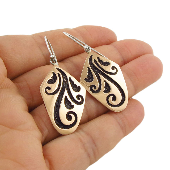 Large mixed metal 925 silver and copper earrings with black floral patterns held in a hand against a white background
