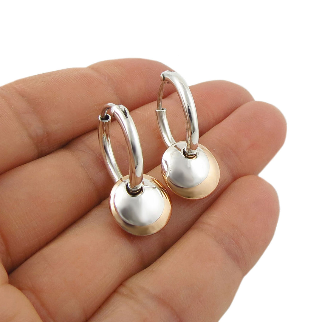 A pair of small hoop earrings with polished 925 sterling silver hoops and round dangling copper discs and pendants, shown resting on an open hand against a white background.