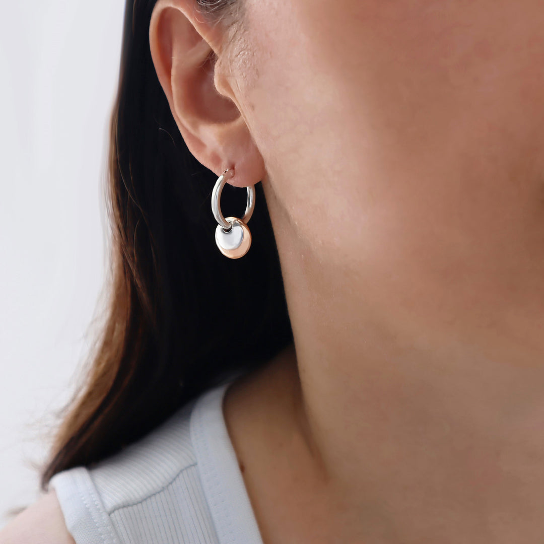 A close-up of a woman's ear wearing a minimalist 925 silver hoop earring with a small hanging copper and silver disc charm, against a soft light background.