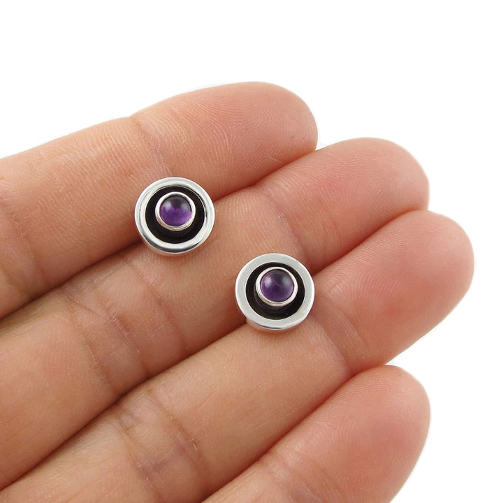Pair of 925 silver stud earrings with purple amethyst birthstone gemstones held between fingers on a white background