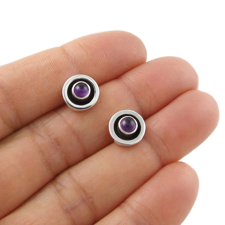 Ladies sterling 925 silver stud earrings with purple amethyst gemstones held between fingers on a white background