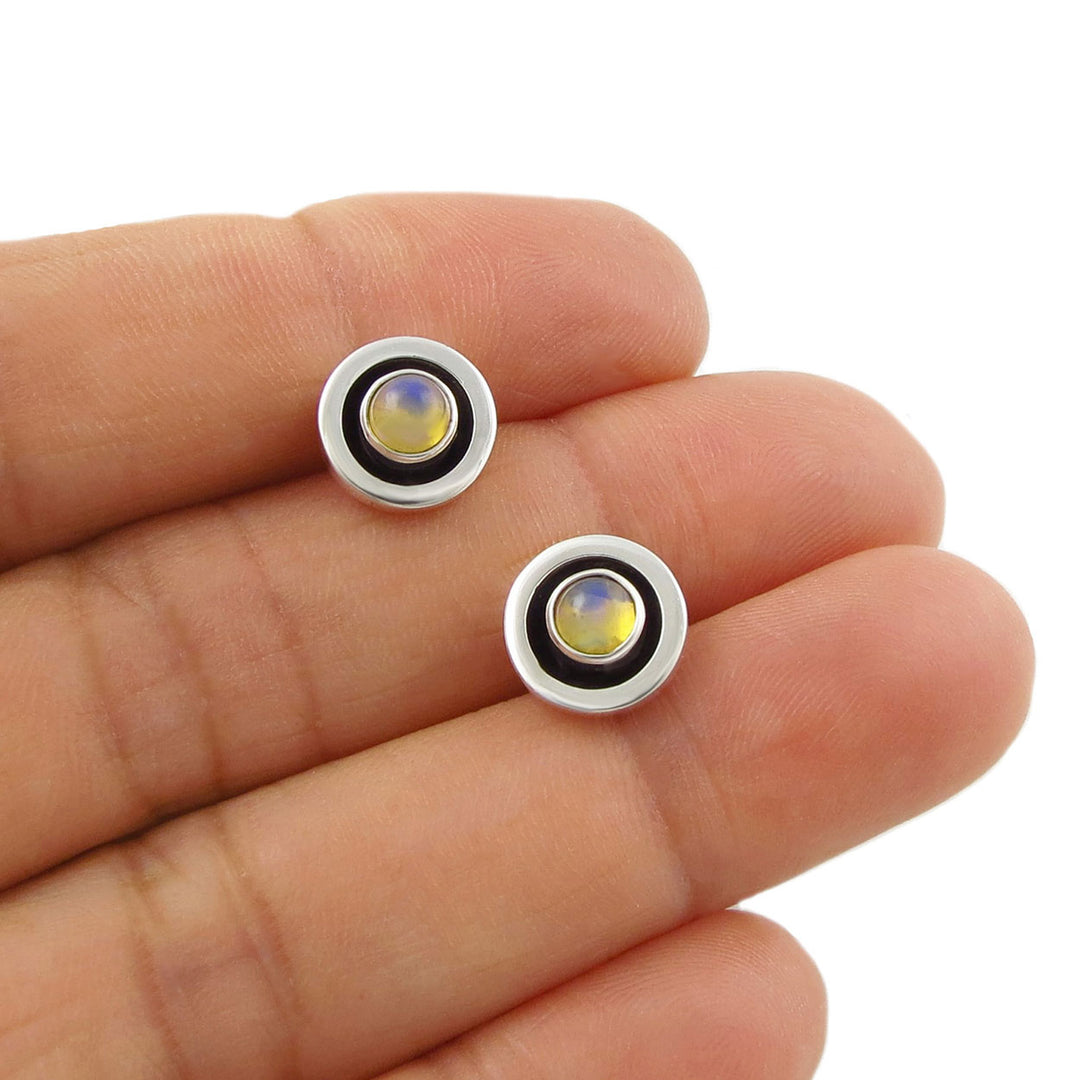 A pair of round sterling 925 silver stud earrings with bezel settings and iridescent yellow real opal gemstones, shown resting on a person's fingers against a white background.