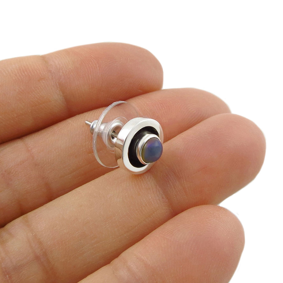 A small sterling 925 silver stud earring with a round dark blue-gray natural opal cabochon stone, shown resting on a person's fingers against a white background.