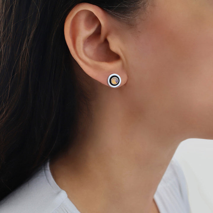 A close-up of a person's ear wearing a small circular 925 sterling silver stud earring with concentric rings in white, black, and a light blue-gray real opal gemstone centre.