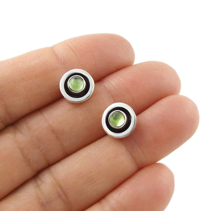Two small round earrings with natural green peridot August birthstone gemstones on a womann's hand.