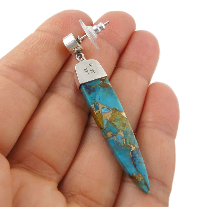 Long turquoise stone earring with 925 sterling silver held between a woman's fingers on a white background