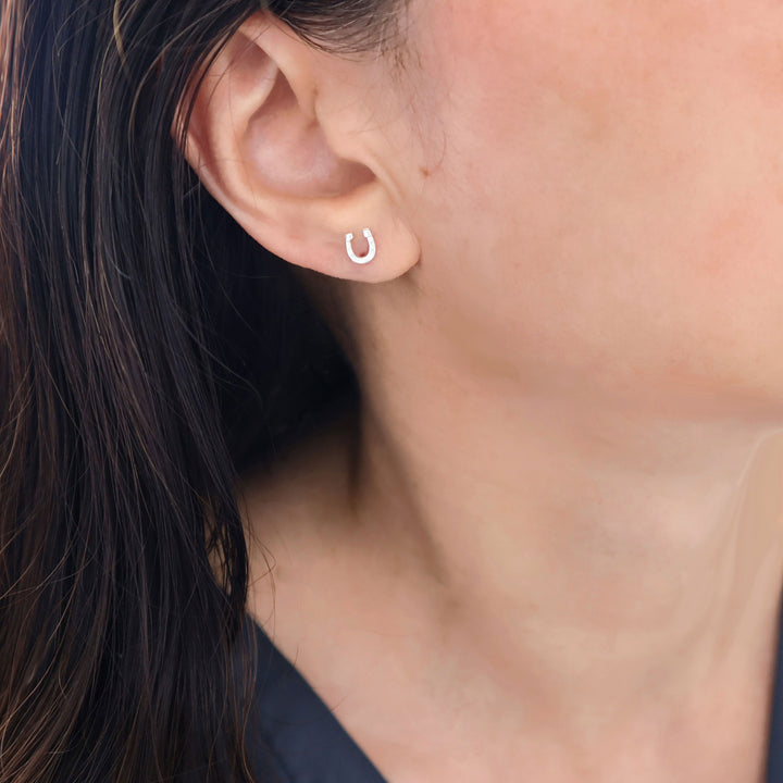 A close-up of a woman's ear wearing a small, minimalist 925 silver crescent-shaped horseshoe stud earring.