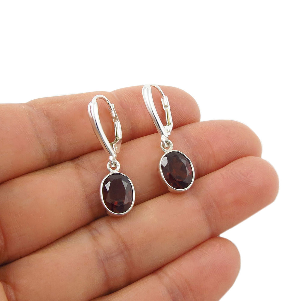 A pair of sterling 925 silver lever-back earrings with oval dark red faceted garnet gemstones, shown resting on a person's hand against a white background.