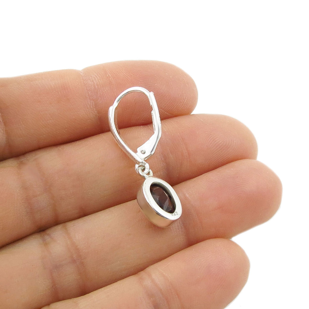 A sterling silver lever-back earring with an oval dark red faceted garnet gemstone, shown resting on a person's fingers against a white background.